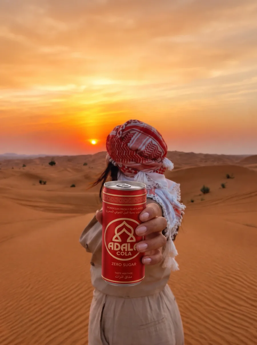 Ambassadrice met een rood-witte keffiyeh in de woestijn bij zonsondergang, die een blikje Adala Cola vooruitsteekt